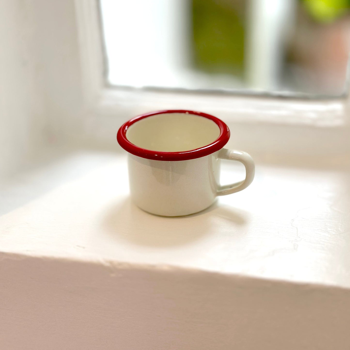 Small white cup with red rim on a light surface near a window
