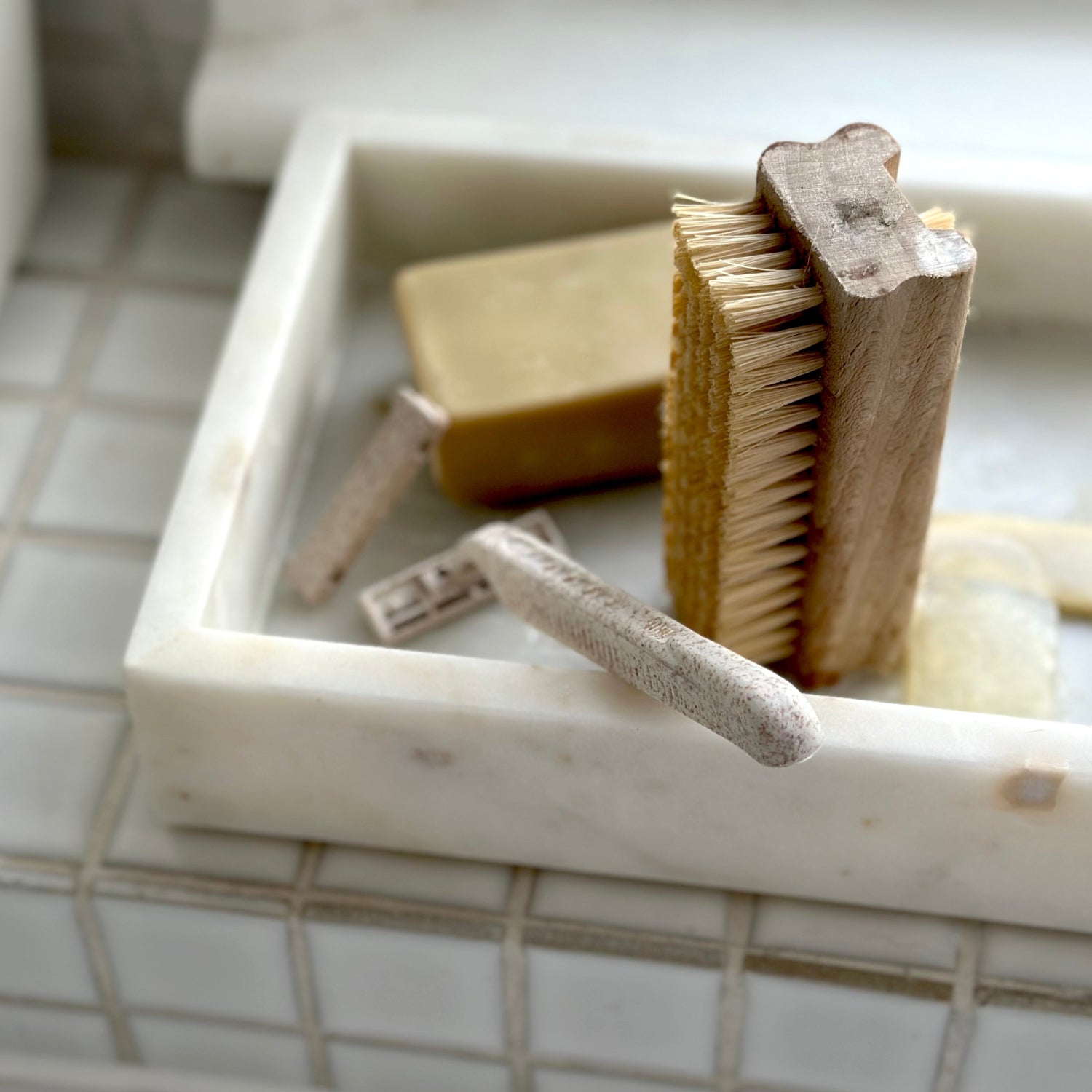 Marble soap dish with various bath items including a brush and soap on a tiled bathroom floor.