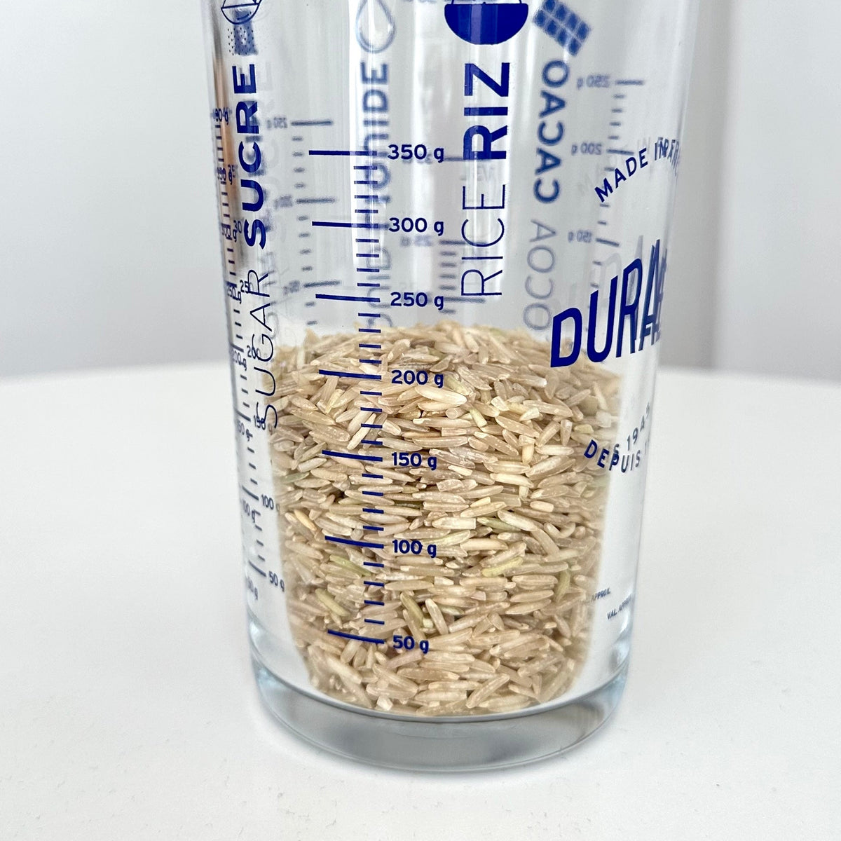 A clear glass measuring cup with blue printed measurement markings and the words 'RICE RIZ' and 'DURALEX' on the side, filled with light tan rice grains, set on a white surface against a white wall.