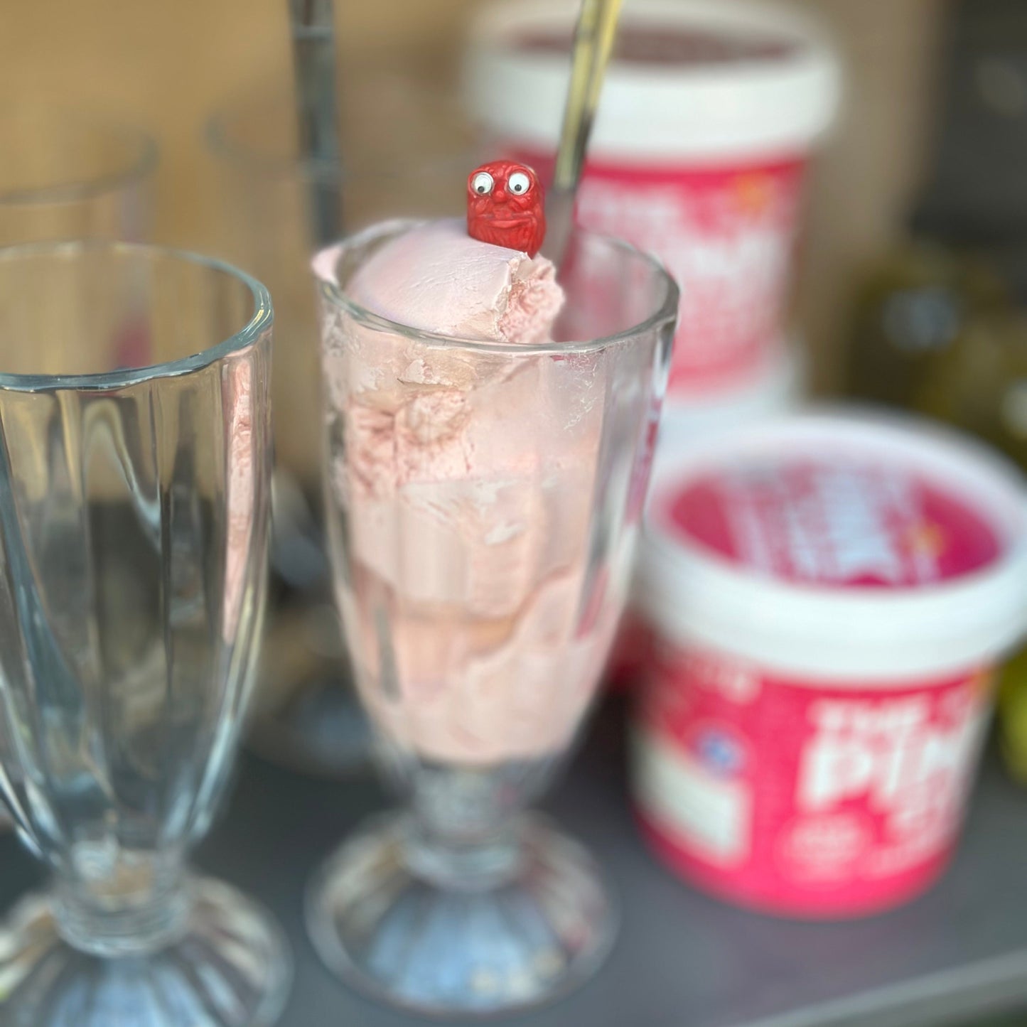 Ice cream in a tall glass with a small red character on top, next to two containers of ice cream.