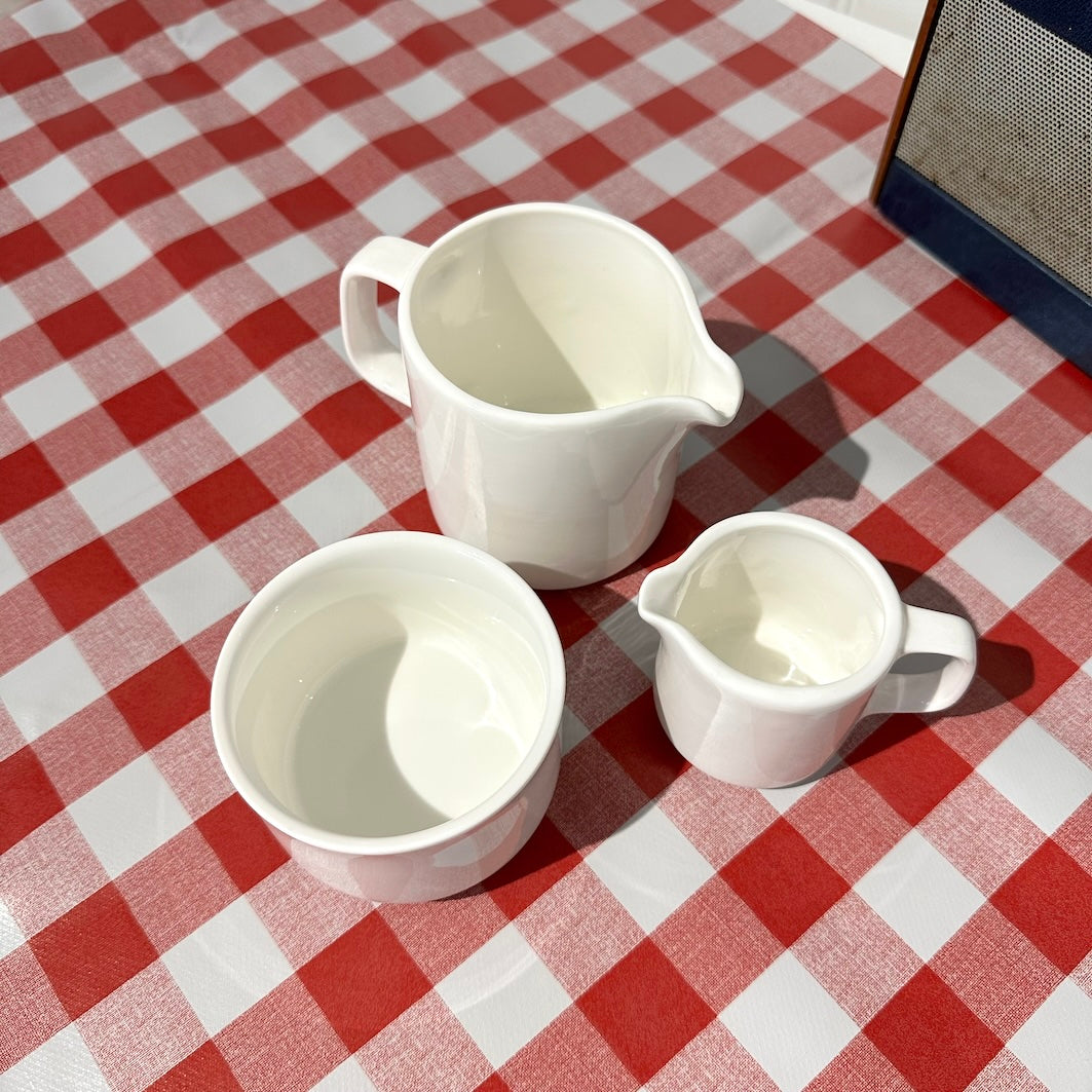 Three white ceramic pitchers on a red and white checkered tablecloth.