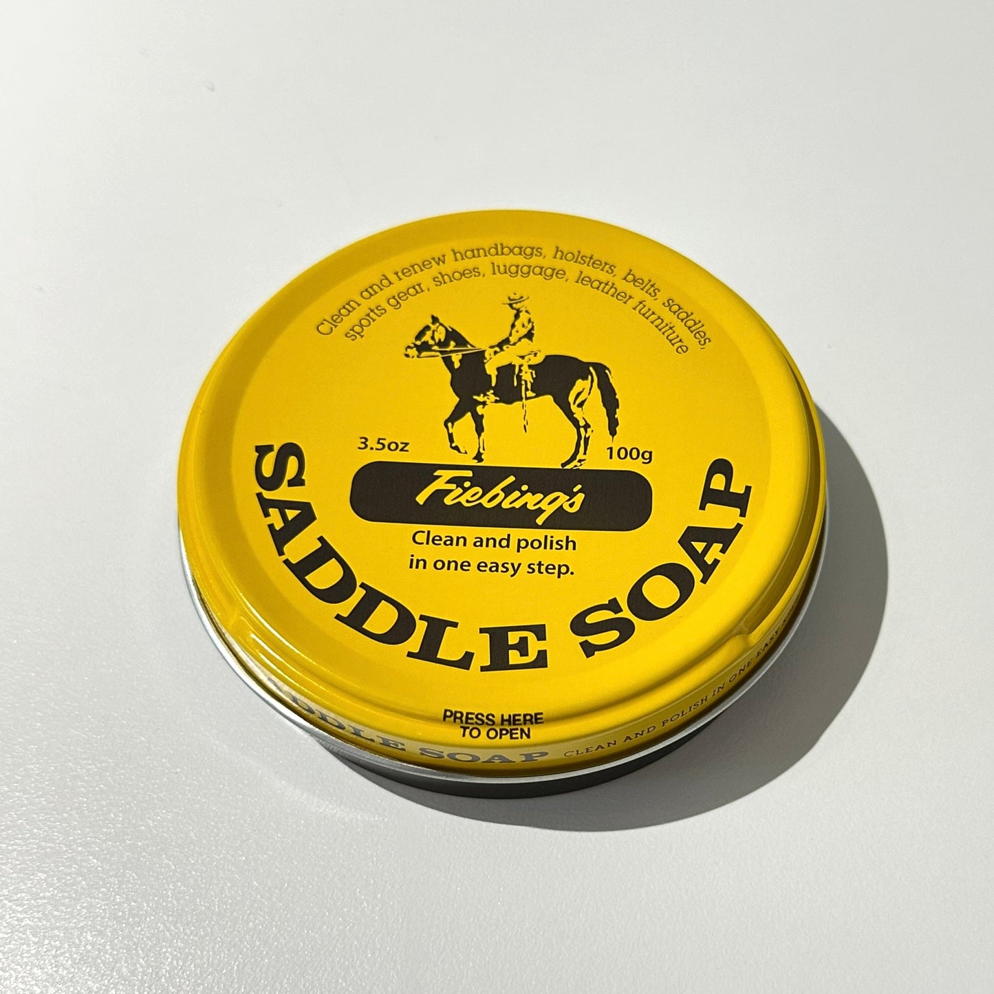 A round yellow tin of Fiebing's Saddle Soap shown on a plain white surface with a soft shadow.