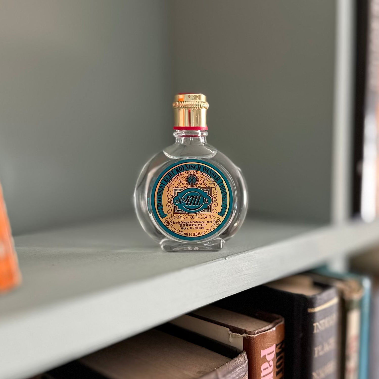A small clear glass bottle  of 4711 cologne with a gold cap and a round blue-and-gold label sits on a gray bookshelf among books.