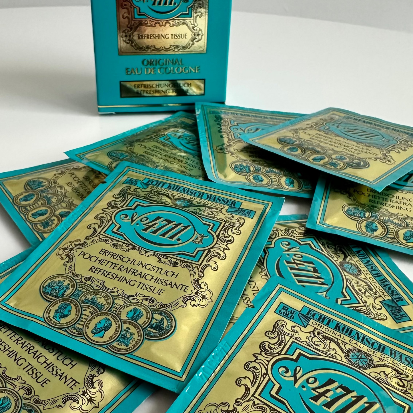 Several small teal and gold cosmetic sachets are scattered on a white surface, with one matching rectangular box standing upright behind them. 