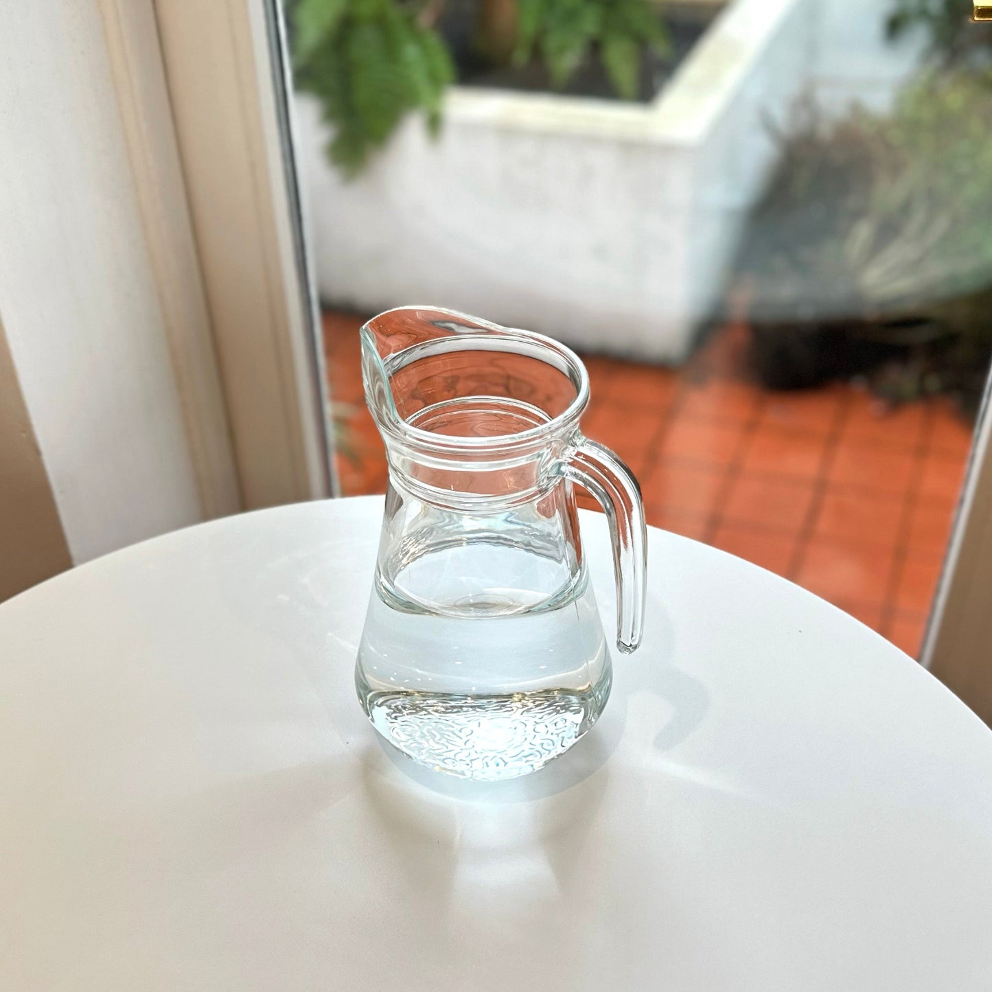 Clear glass pitcher with water on a white surface, blurred indoor background