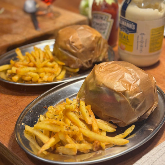 Two burgers and fries on metal trays with condiments in the background.