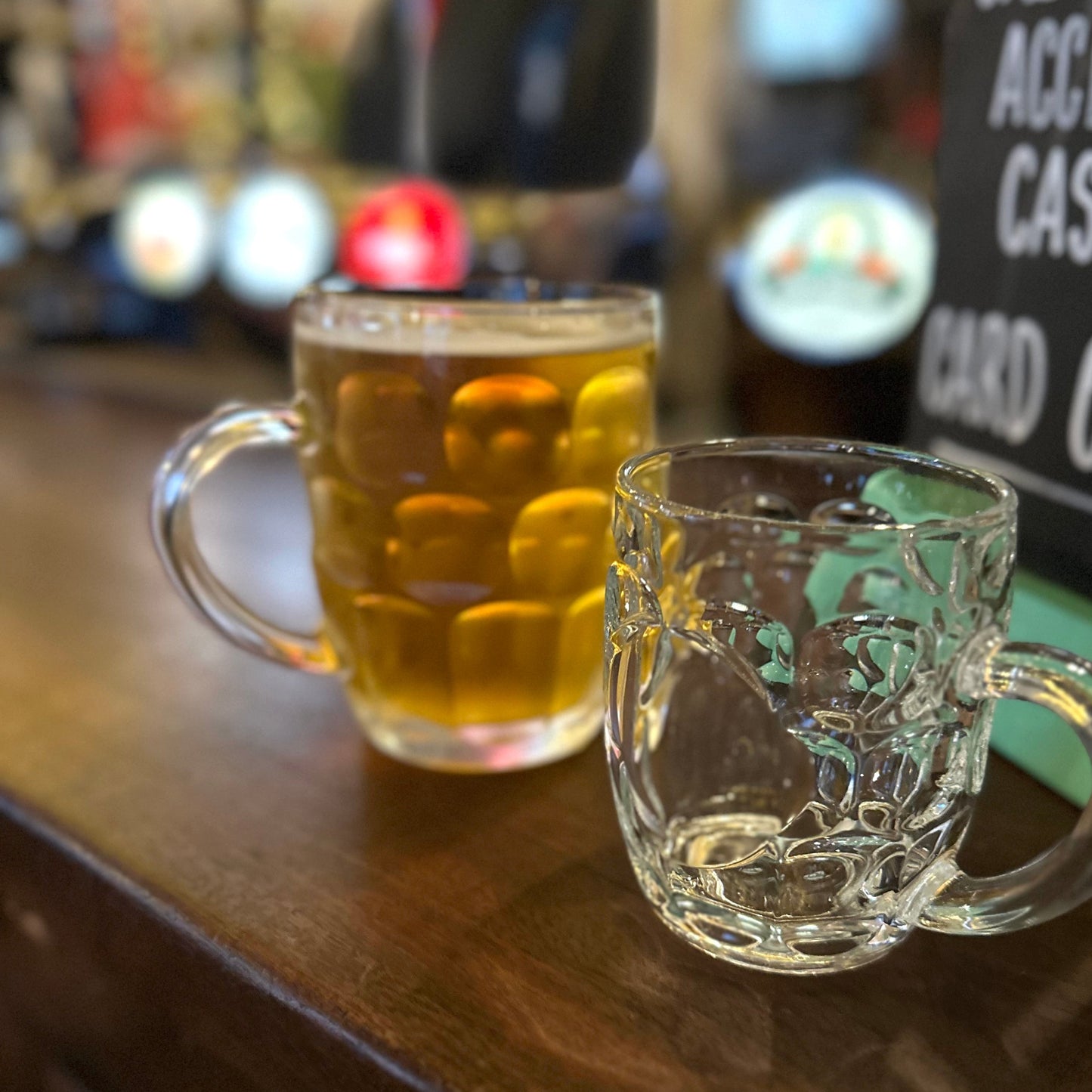 Two glass mugs on a bar counter with one containing a drink, blurred background with 'We Do Accept Cash' sign.