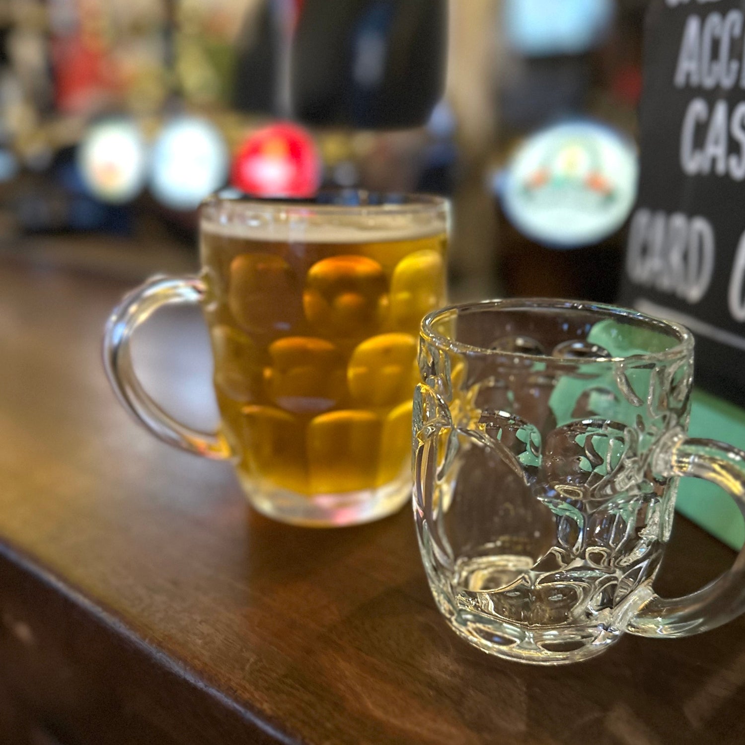 Two glass mugs on a bar counter with one containing a drink, blurred background with 'We Do Accept Cash' sign.