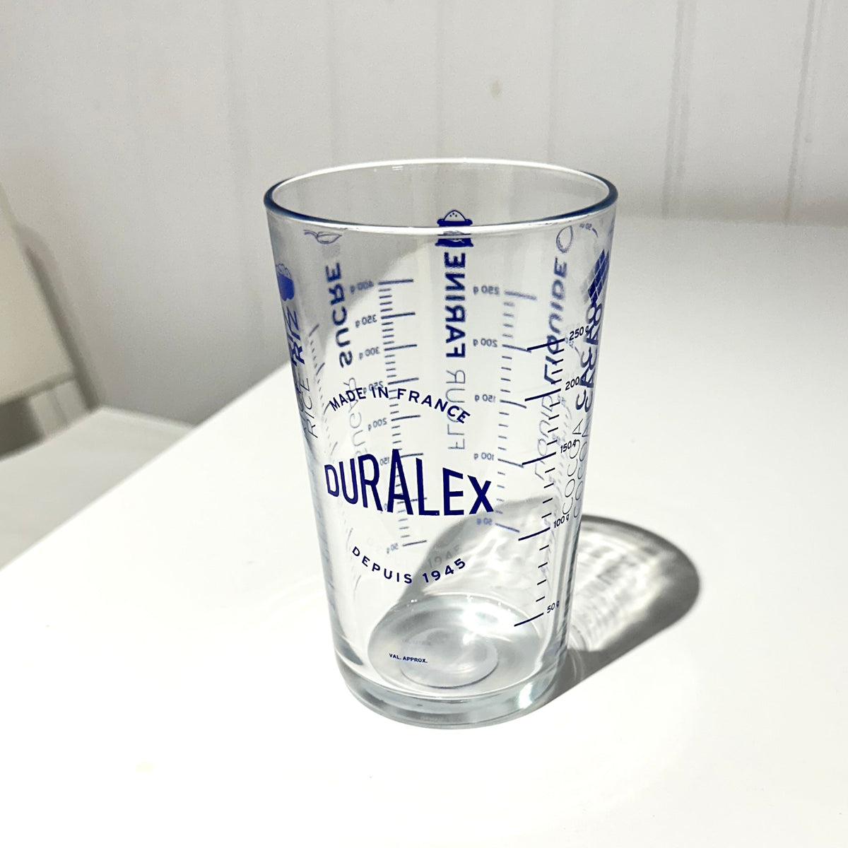 A clear glass measuring tumbler with blue printed measurement markings and the brand name Duralex, sitting on a white table against a white panelled wall.