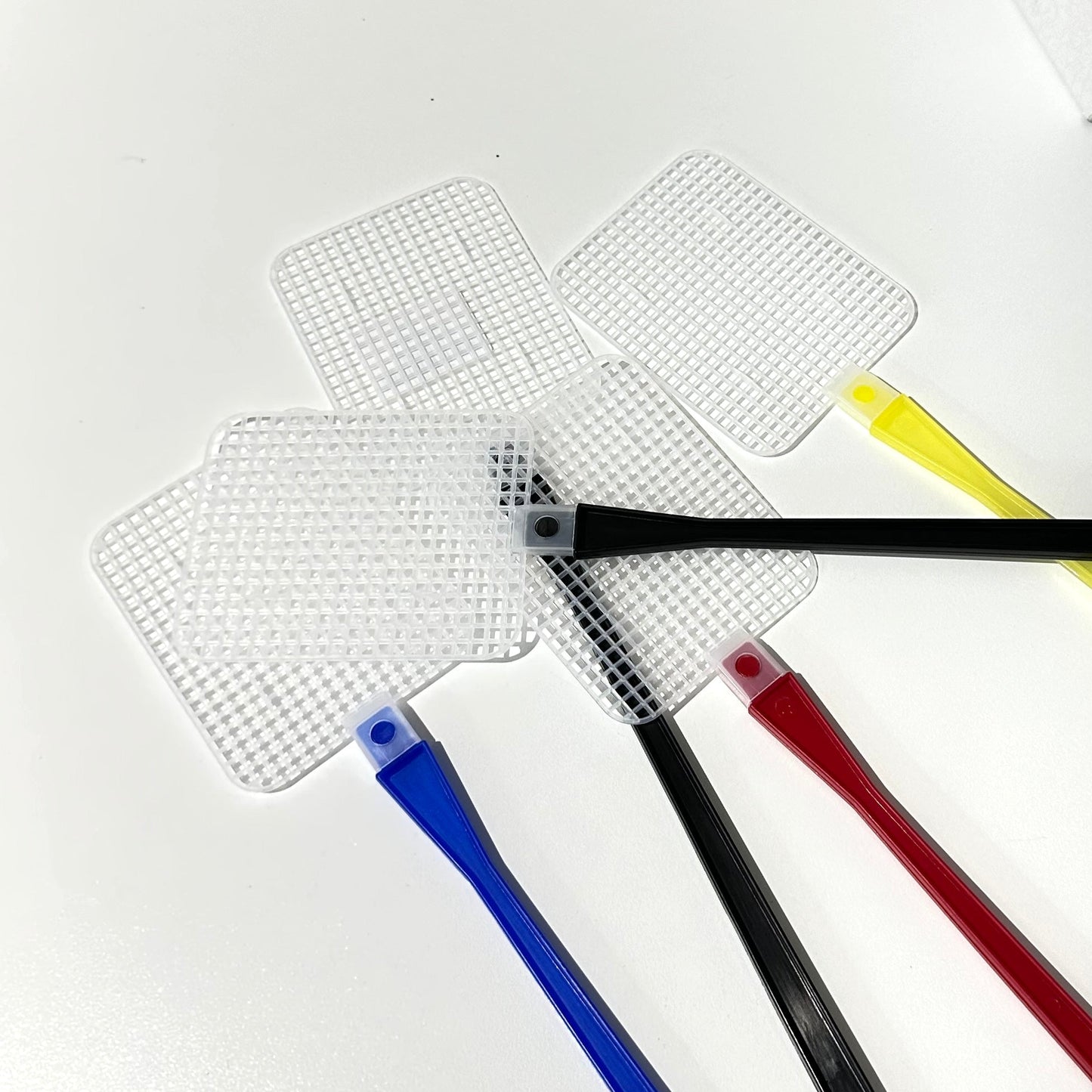 Four fly swatters with colored handles in blue, black, red, and yellow, laid out on a white table next to their white branded packaging box.