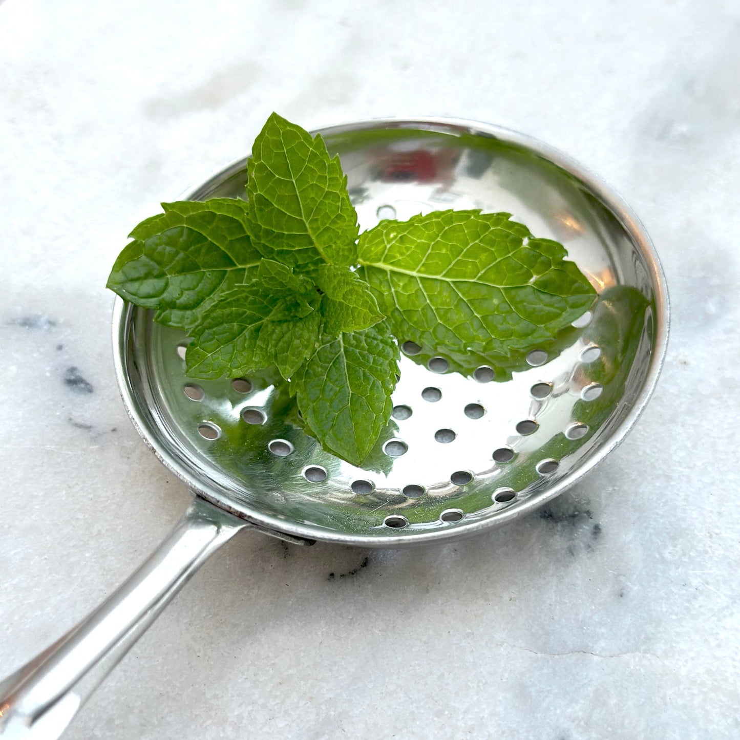 Metallic strainer with mint leaves on a marble surface
