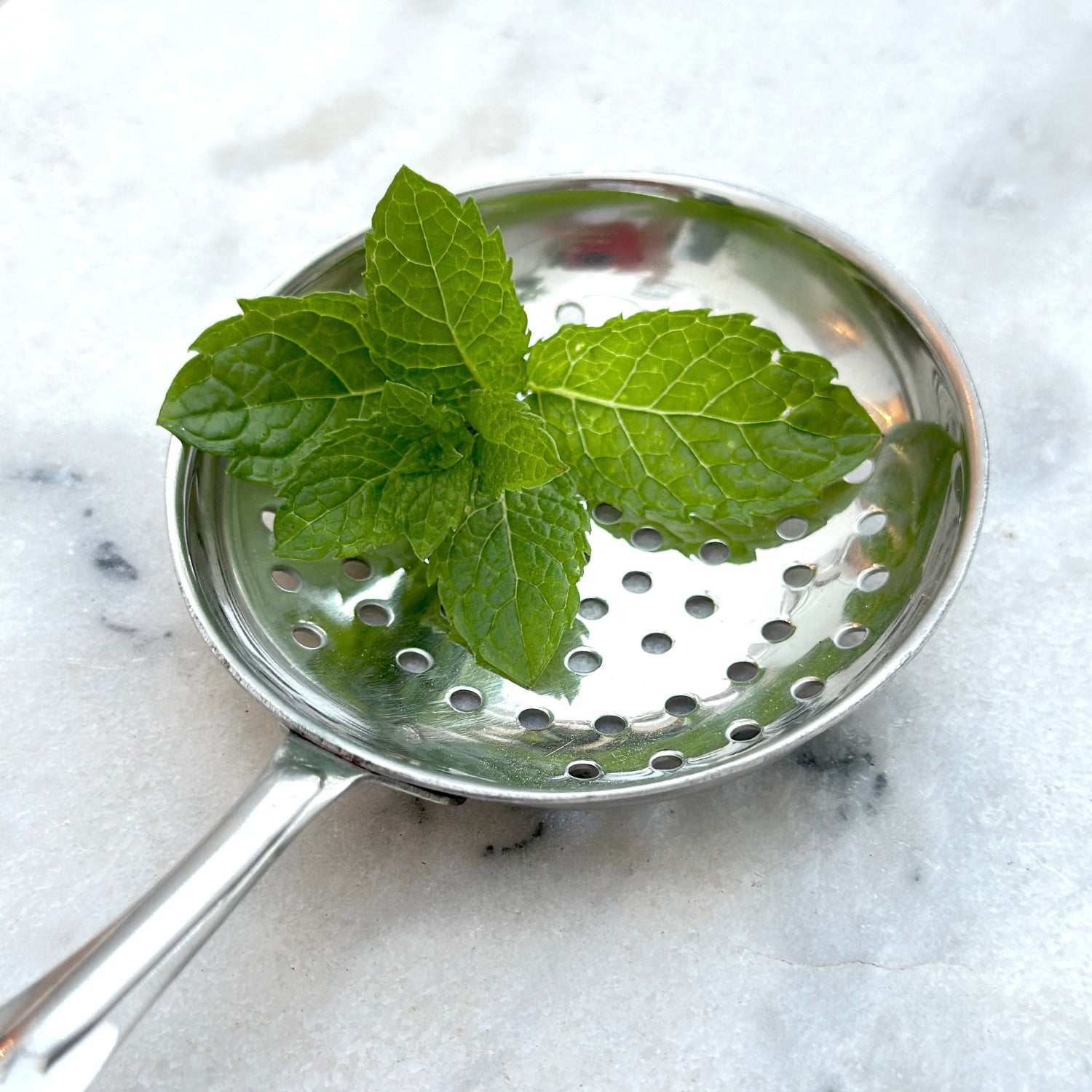Metallic strainer with mint leaves on a marble surface