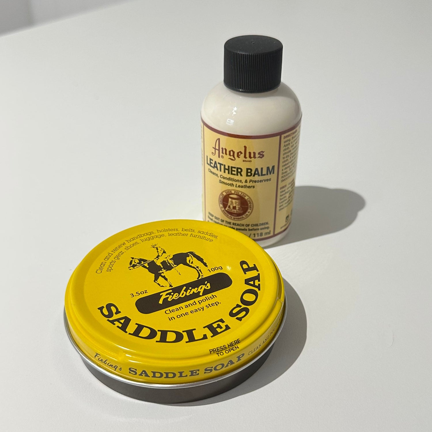 A small bottle of Angelus Leather Balm and a round tin of Angelus Saddle Soap are displayed on a white surface against a white paneled wall.