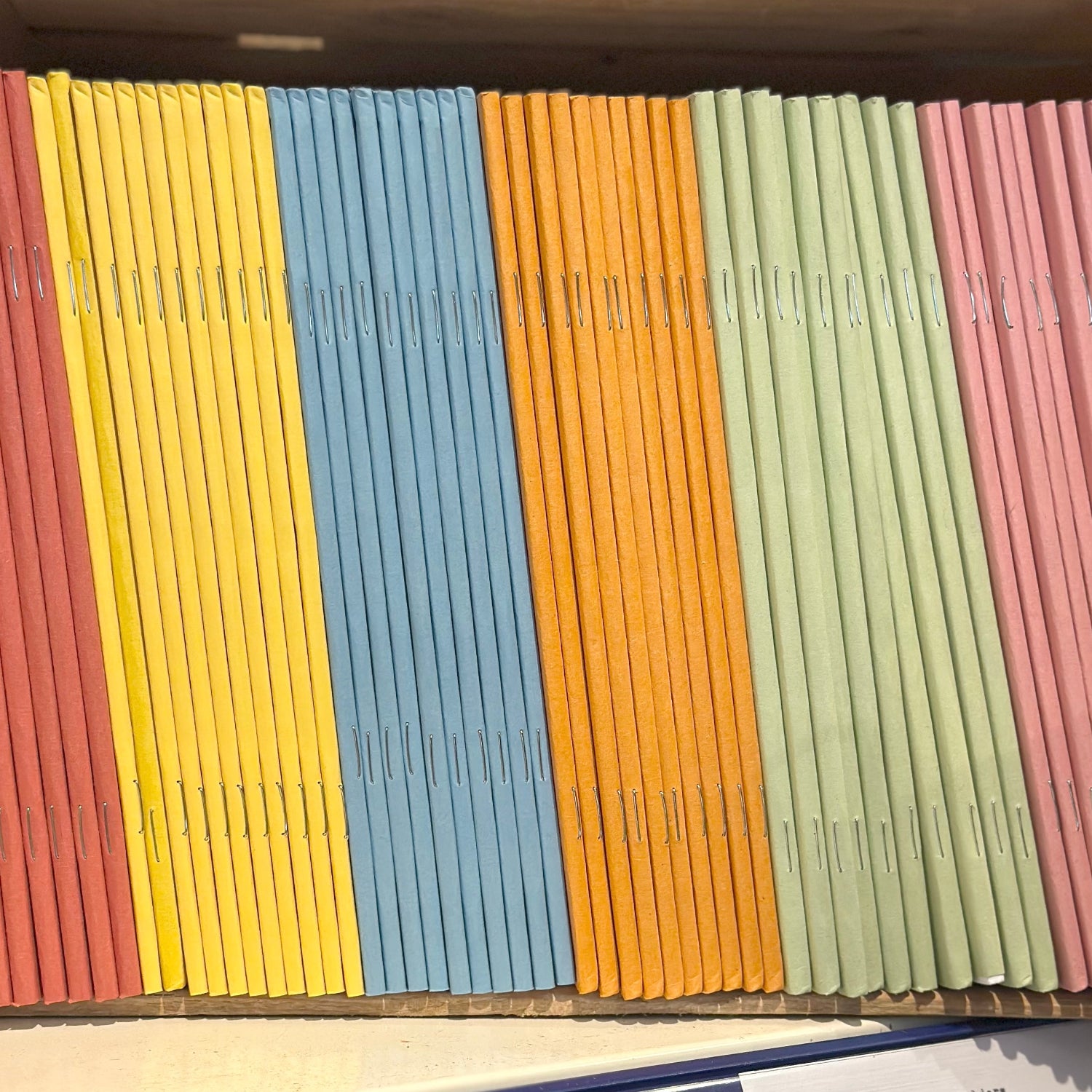 Coloured exercise books arranged in a box with labels on a shelf.