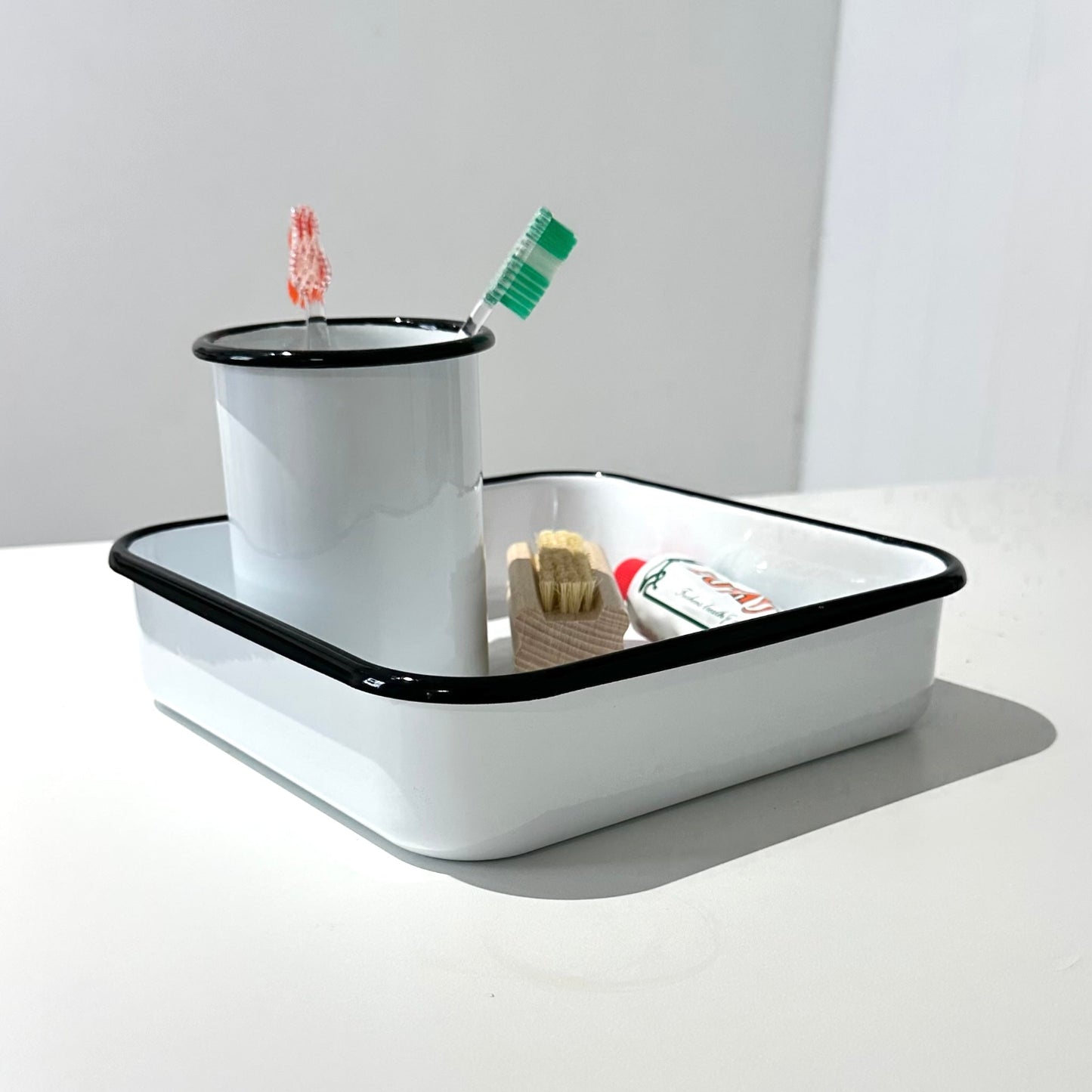 A white enamel toothbrush holder with a black rim sits on a white table in a bright room. A toothbrush with a green handle and a small toothbrush holder insert are placed inside the tray.