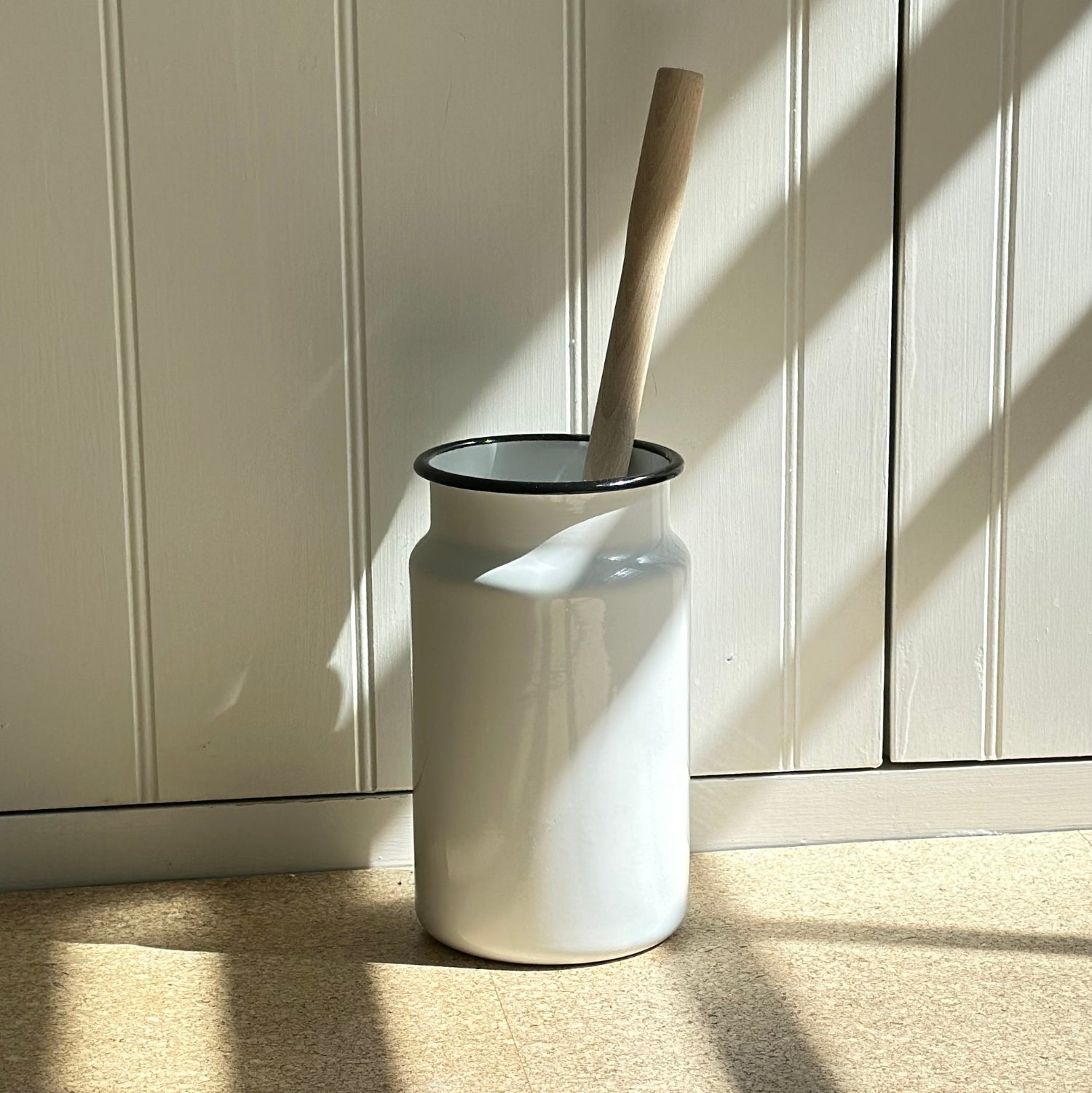 White enamel toilet brush holder with a wooden  brush against a light wooden panel wall.