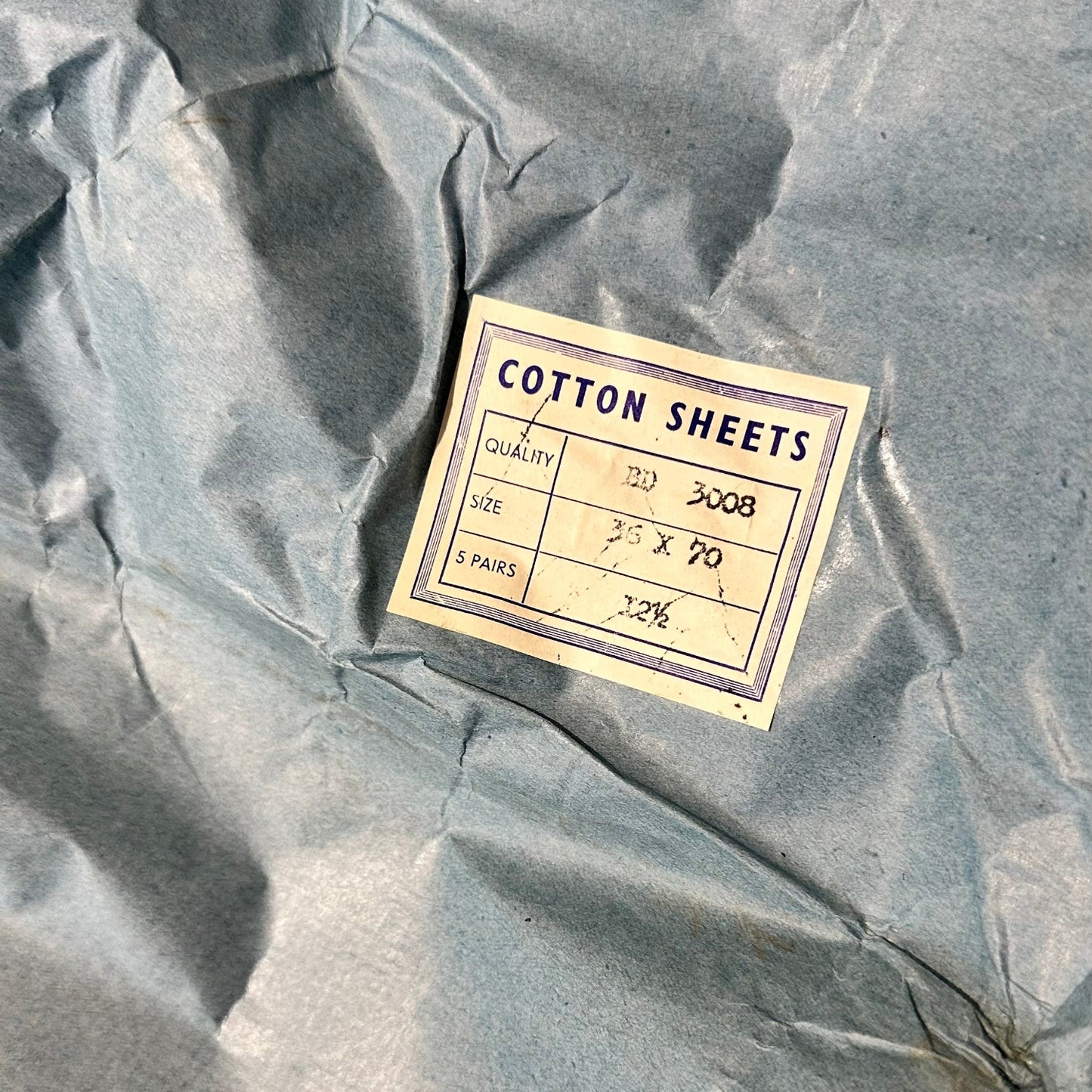 A close-up of crumpled gray-blue tissue paper with a small cream paper label reading 'COTTON SHEETS' and handwritten details.